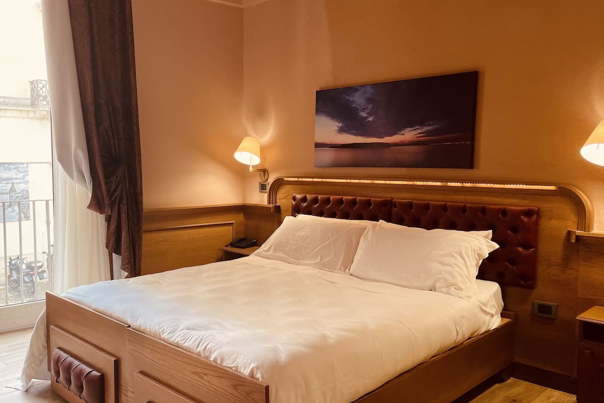 A hotel room with a large bed, wooden headboard, two bedside lamps, a painting on the wall, and a balcony with a view of the city.