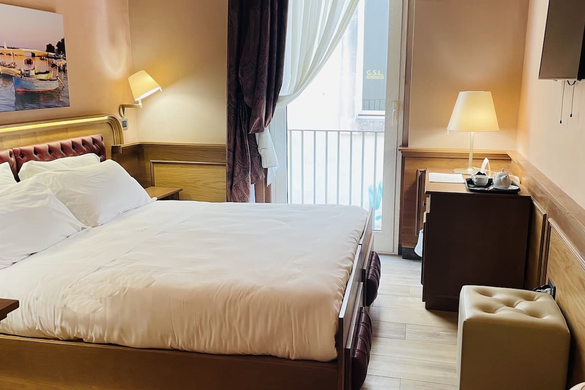 A hotel room with a bed, bedside table, lamp, and a view of the outside.