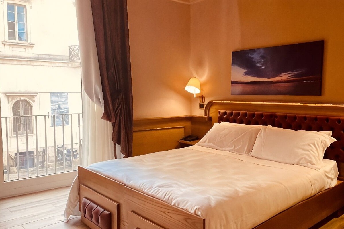 Ein Hotelzimmer mit einem Bett, zwei Nachttischlampen, einem Bild an der Wand und Blick ins Freie.