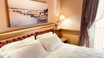 Una camera d'albergo con un letto, un comodino, una lampada, un quadro raffigurante una scena sul mare e una finestra con tende.