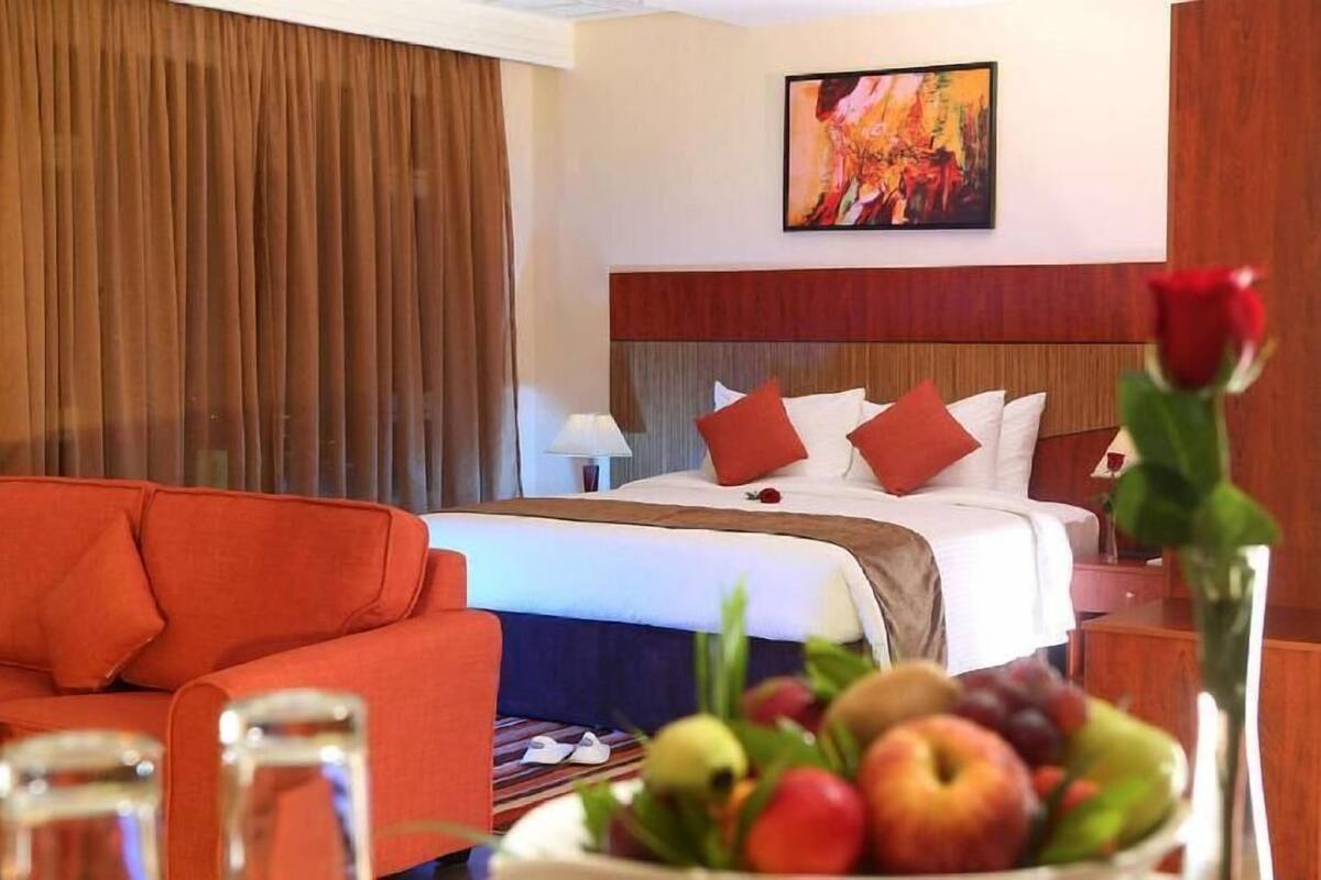 A hotel room with a bed, a red sofa, a fruit bowl, and a painting on the wall.