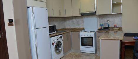 A compact kitchen with a refrigerator, washing machine, microwave, stove, and tiled backsplash.