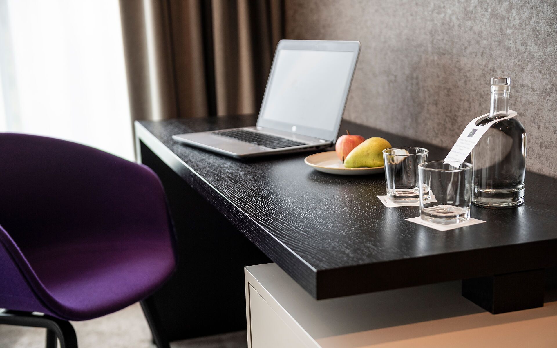 Una laptop sobre un escritorio de madera oscura, una botella, dos vasos y un plato con fruta.