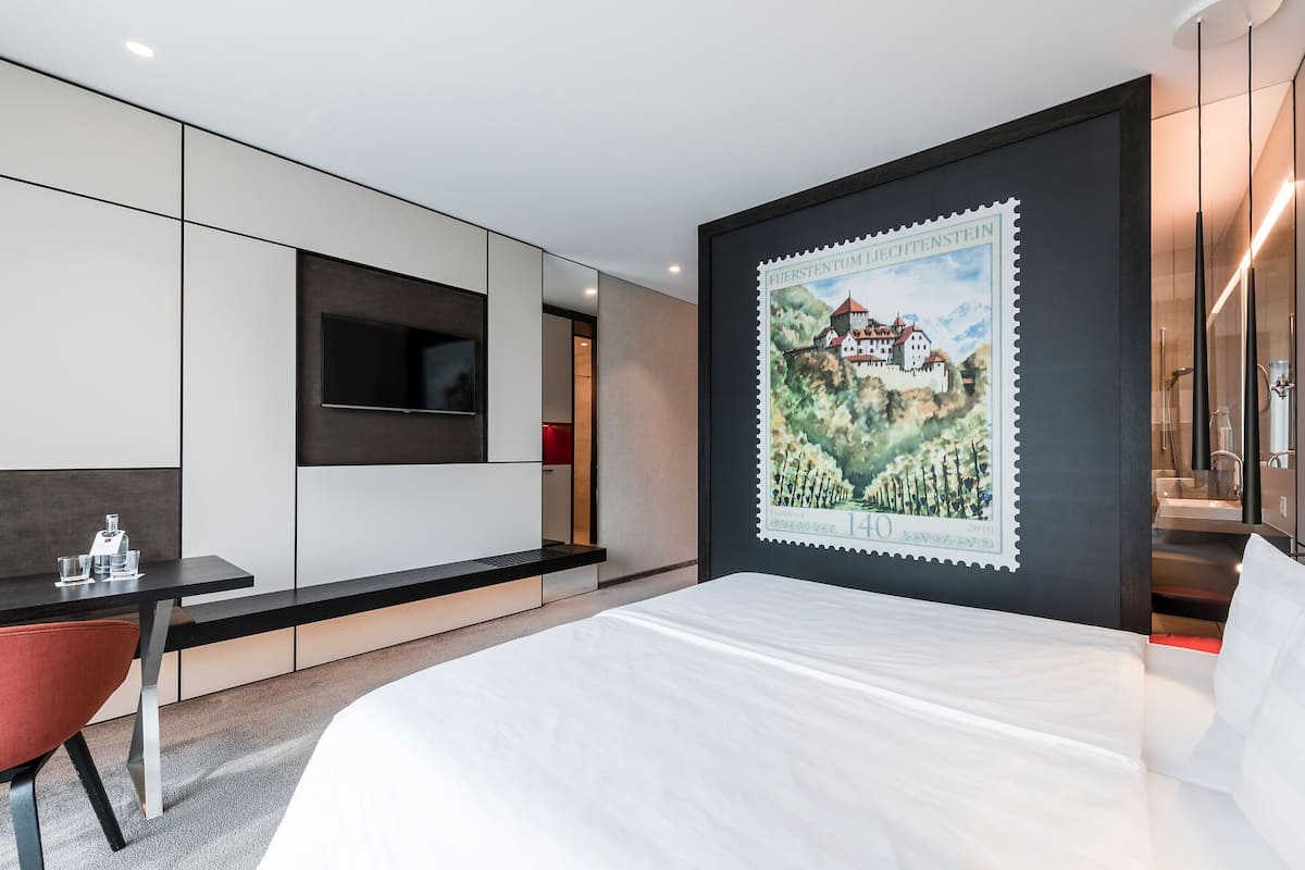 A modern hotel room with a bed, a desk, a TV, and a large poster on the wall.