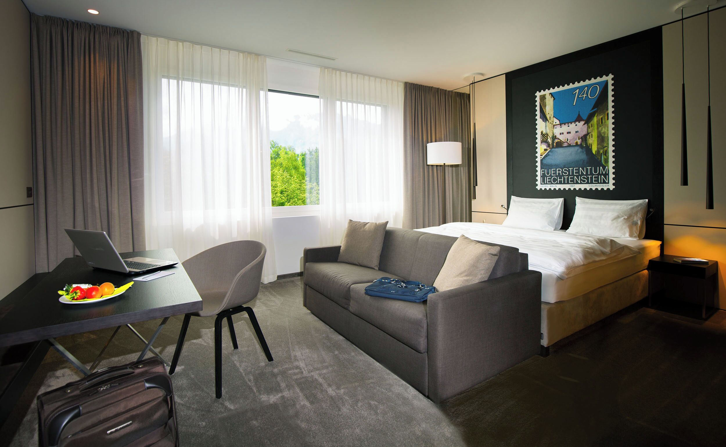 A hotel room with a bed, a desk with a laptop, a chair, and a view of greenery outside.