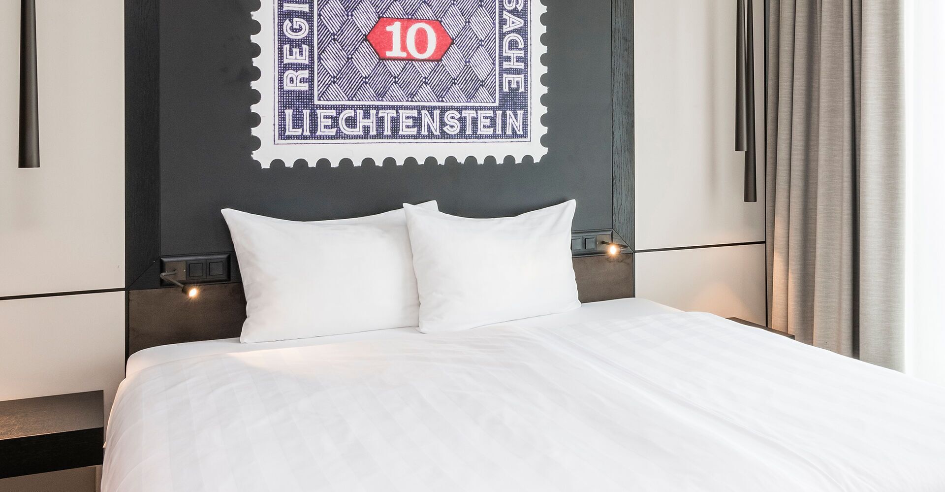 Ein Hotelzimmer mit Bett, Kissen, einer Wandleuchte und einem gerahmten Briefmarkenbild.