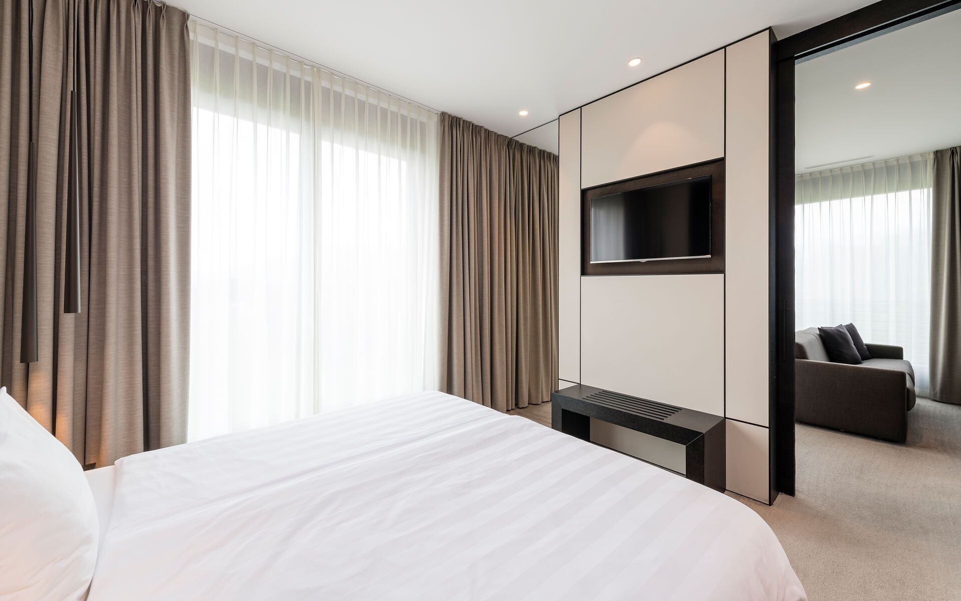 A modern hotel room with a large bed, a flat-screen TV mounted on the wall, and a seating area with a sofa and chairs.