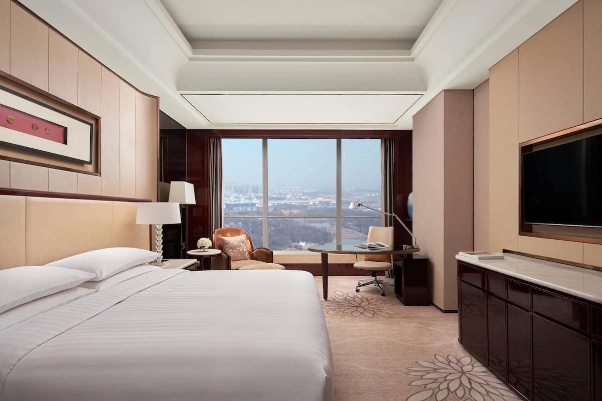 A hotel room with a large bed, a desk with a chair, a TV, and a view of the city through a large window.