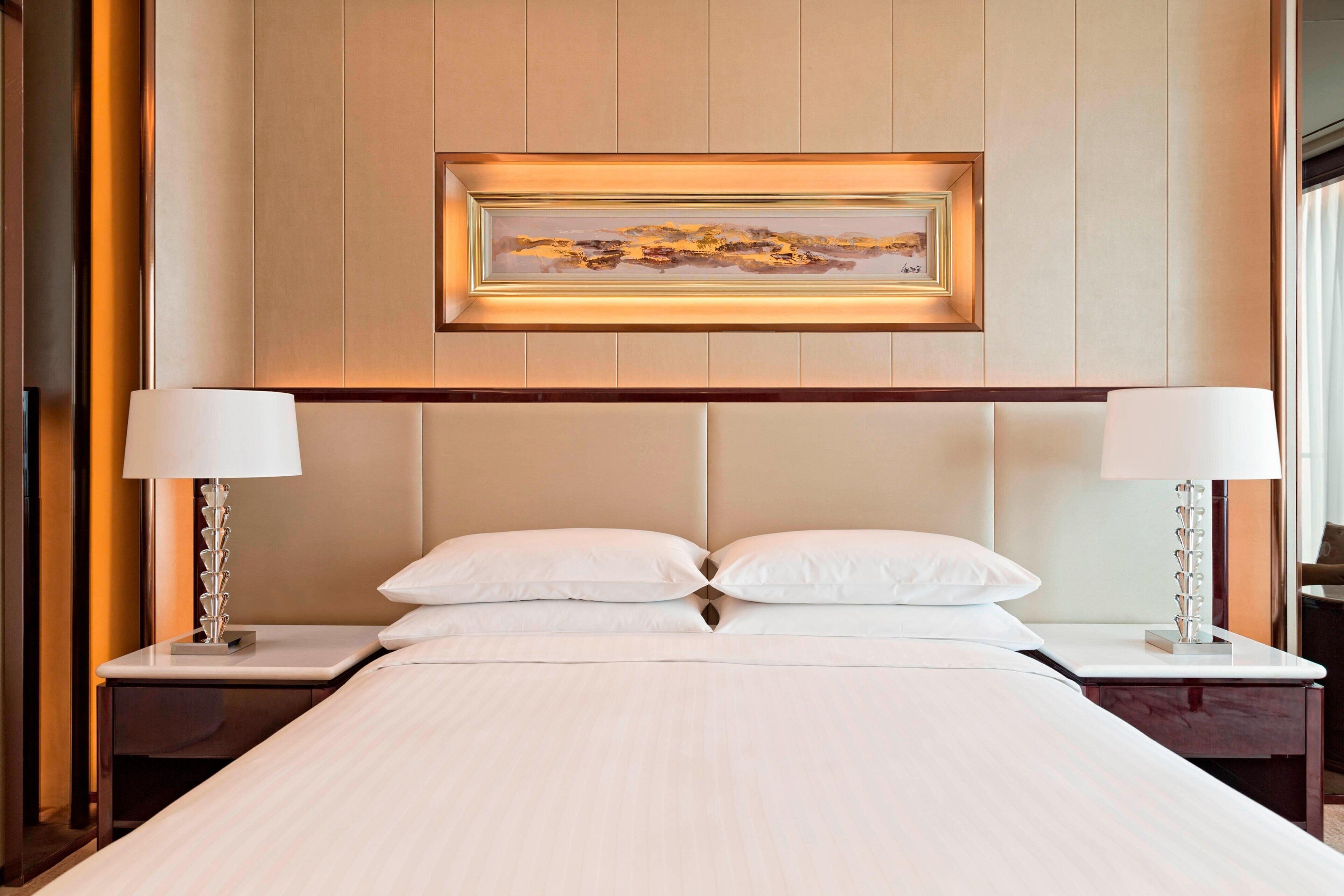 A hotel room with a large bed, two bedside tables with lamps, a framed artwork above the bed, and a wooden headboard.