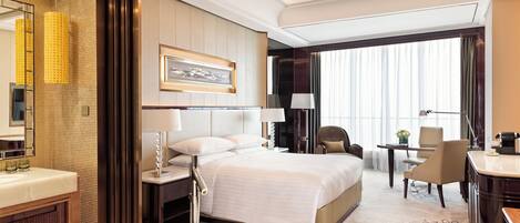 A hotel room with a large bed, a sitting area with a chair and table, a bathroom with a bathtub, and a large window with curtains.