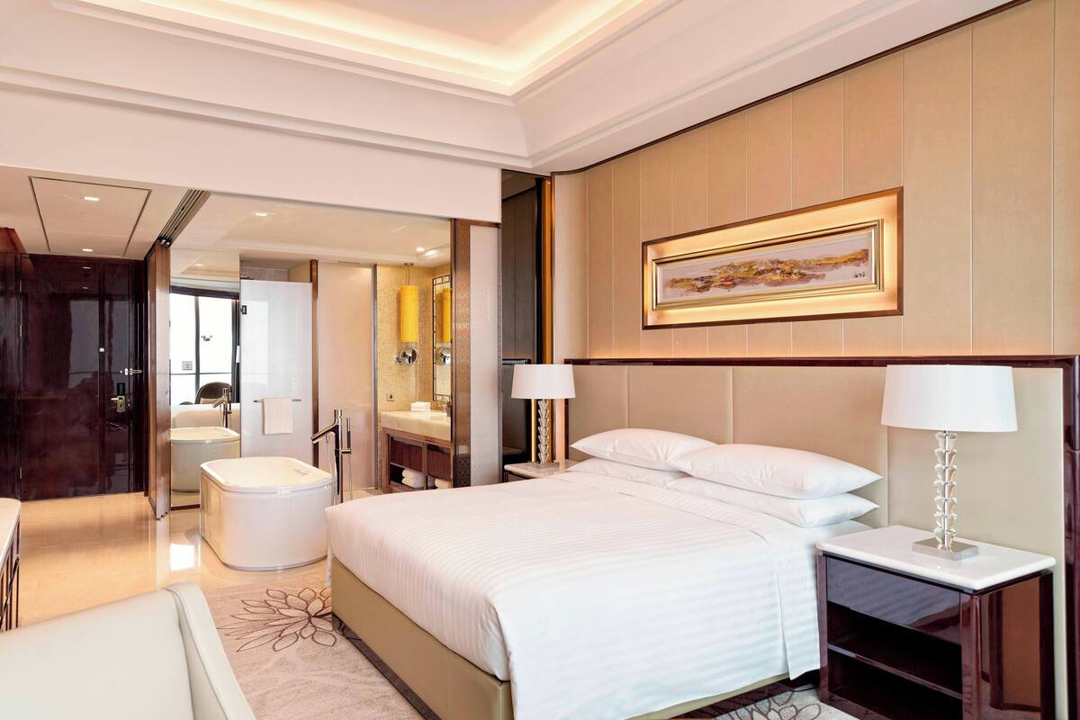 A hotel room with a large bed, a bedside table with a lamp, a bathroom with a bathtub, and a mirror.