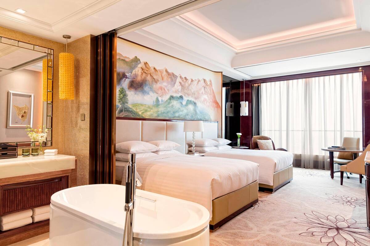 A hotel room with two beds, a large painting, a bathroom with a bathtub, and a desk with a chair.