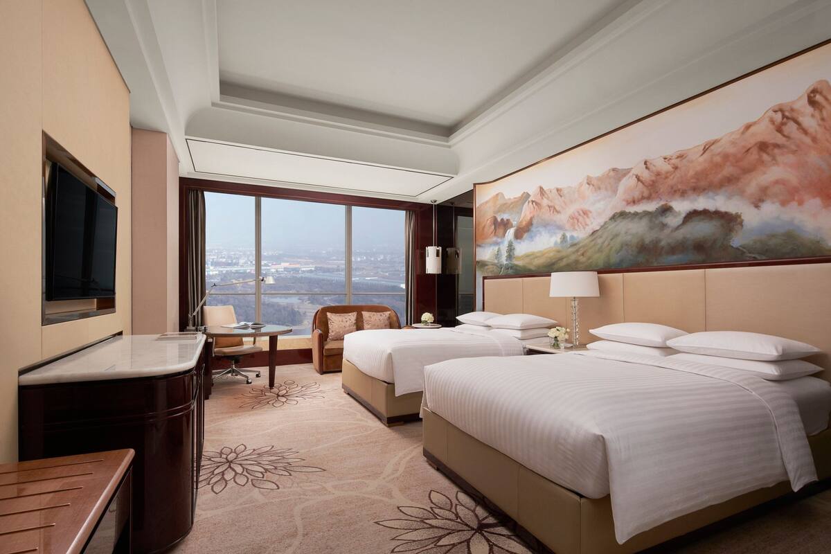 A hotel room with two beds, a desk, a chair, and a large window offering a city view.