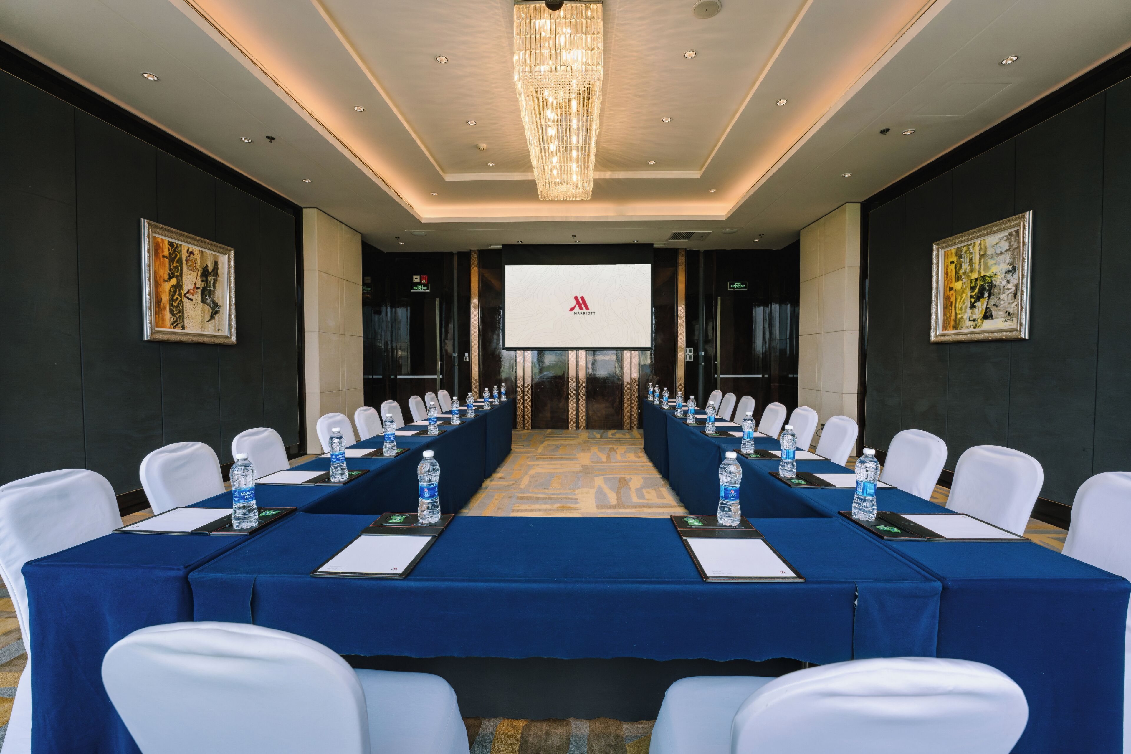A conference room with a long table, white chairs, water bottles, and a projector screen.