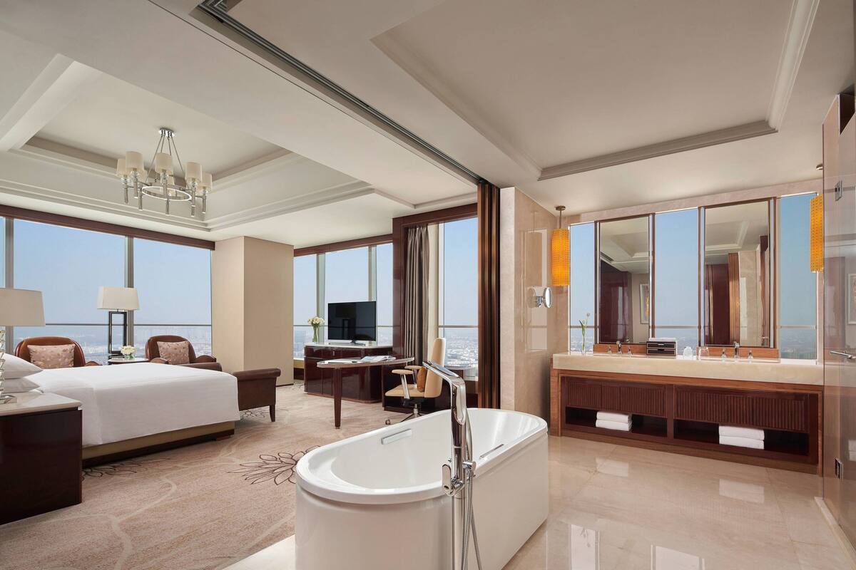 A hotel room with a large bed, a sitting area, a bathtub, a vanity with a mirror, and a television.