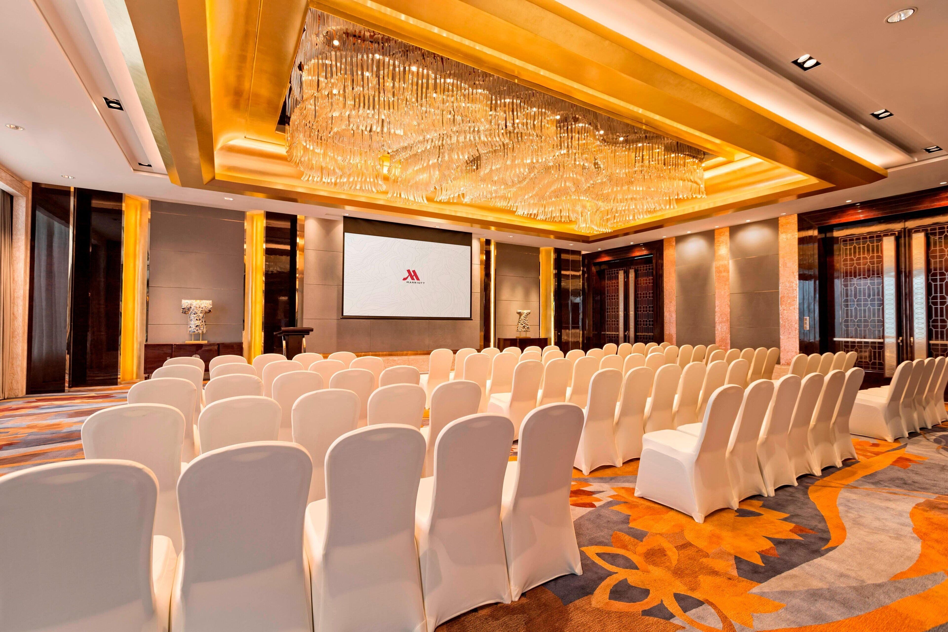 A conference room with rows of white chairs, a large chandelier, and a projection screen.
