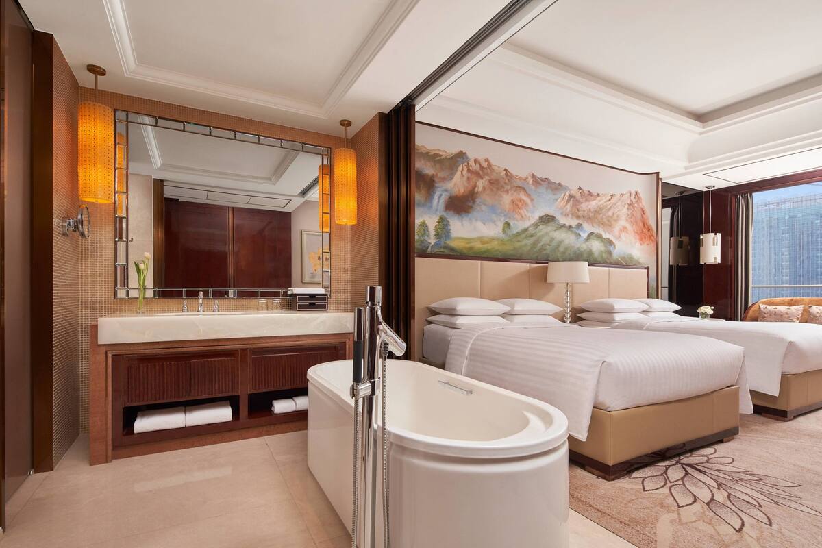 A hotel room with a large bed, a separate bathroom with a bathtub, a large mirror, and a painting of mountains.