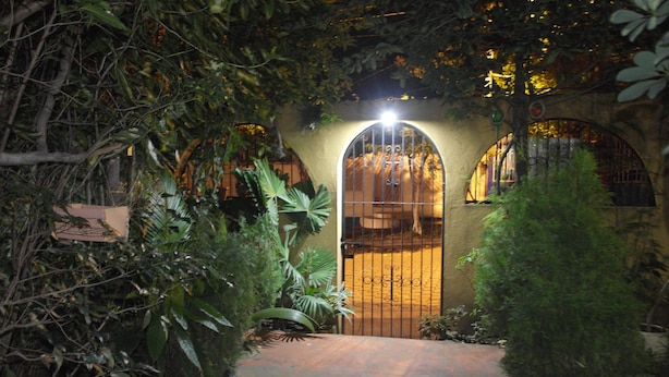 Une entrée en arc surmontée d’un portail en métal, entourée d’une végétation luxuriante et bordée d’un chemin pavé menant à un bâtiment dont le mur est jaune.