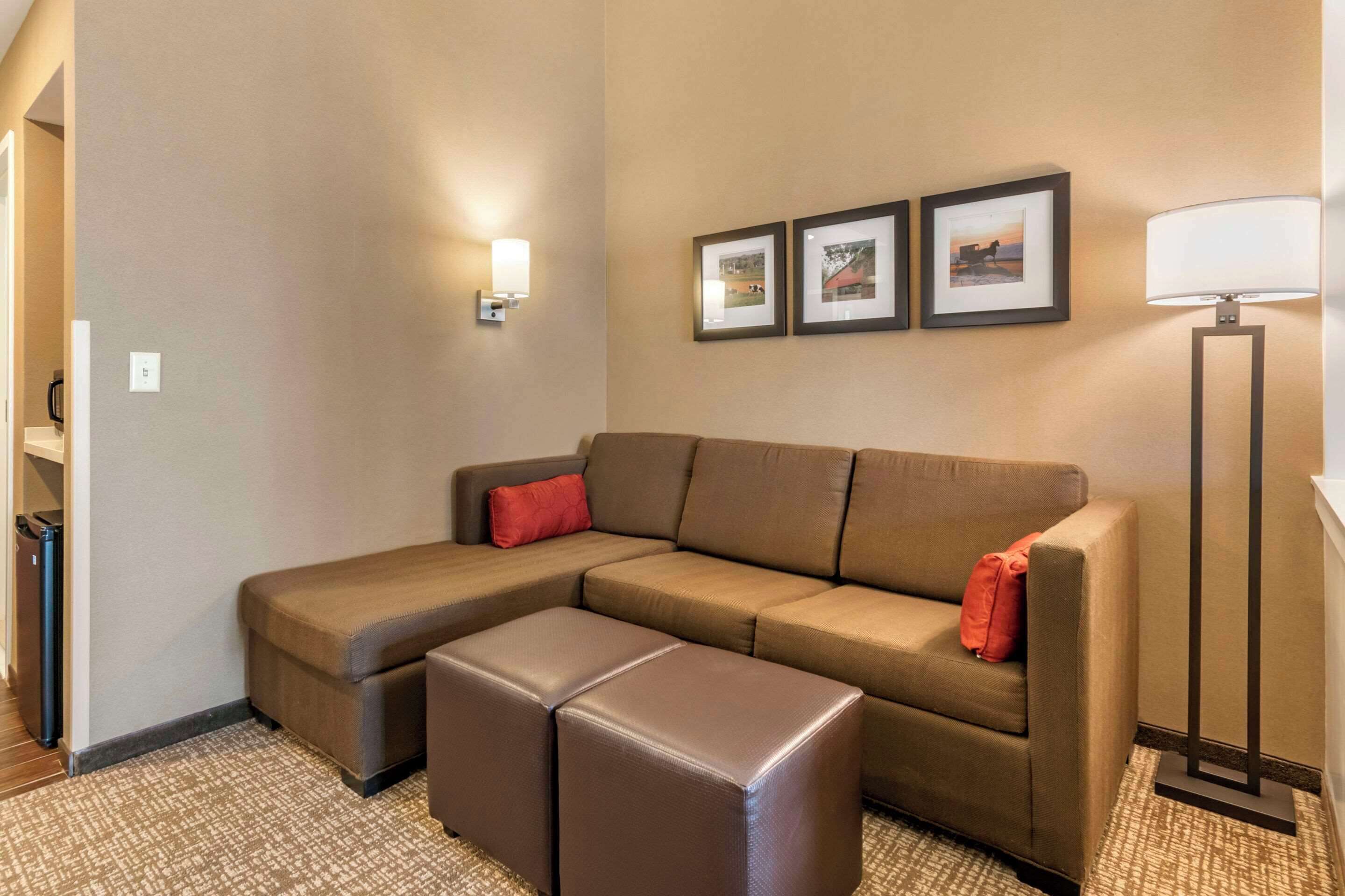 Una habitación de hotel moderna con sofá modular, un pequeño otoman, una lámpara de pie y cuadros en la pared.