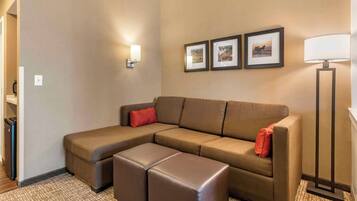 Una habitación de hotel moderna con sofá modular, un pequeño otoman, una lámpara de pie y cuadros en la pared.