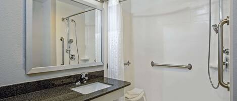 Baño con lavamanos de encimera de granito, espejo, ducha con puerta de vidrio y una toalla blanca sobre un taburete pequeño.