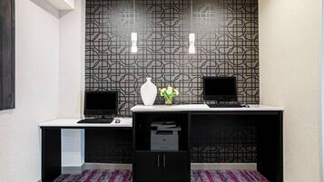 Een moderne kantoorruimte met twee bureaus, elk met een computerbeeldscherm, een witte vaas met bloemen en een printer. De kamer heeft behang met een patroon en een vloerkleed.