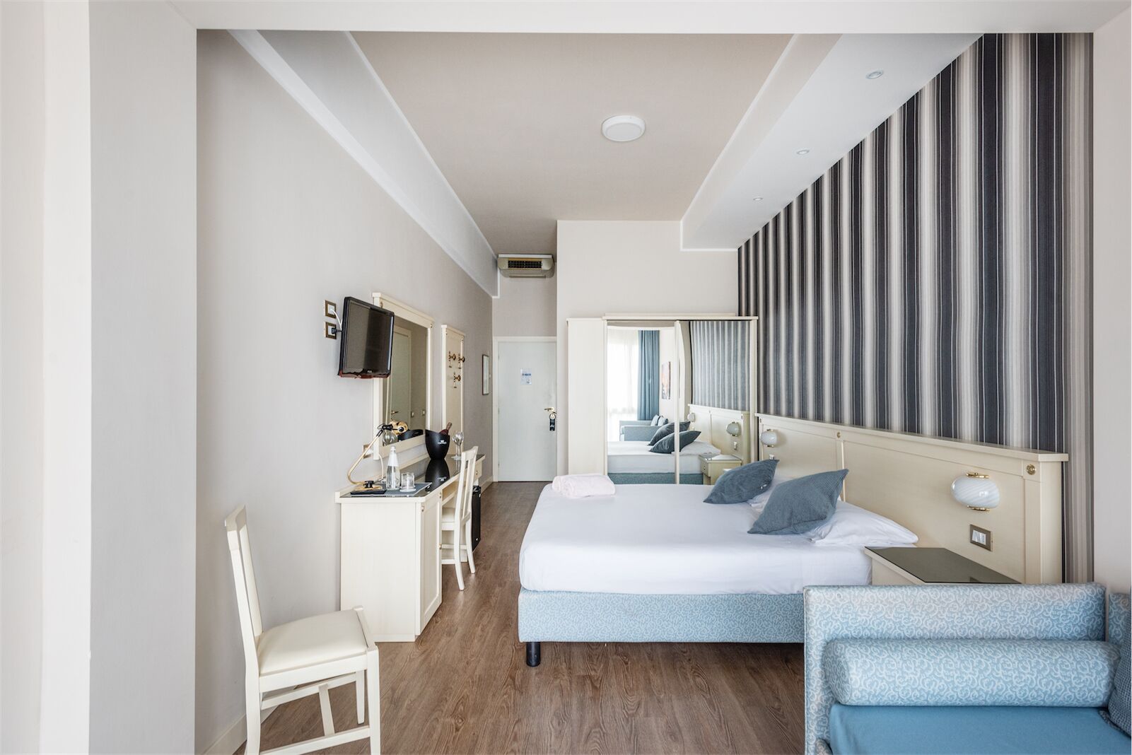 A modern hotel room with a large bed, a desk with a chair, a TV, and a mirror.