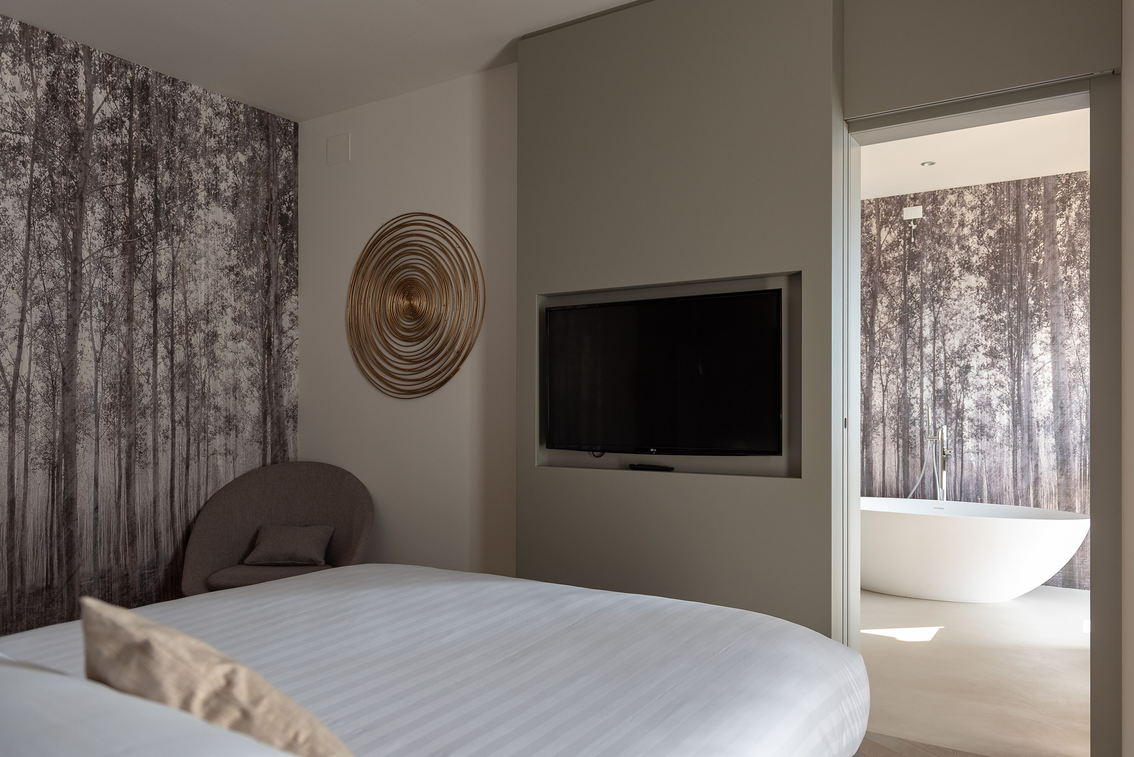 A modern bedroom with a large bed, a television, a circular wall art piece, and a view of a forested area through a window.