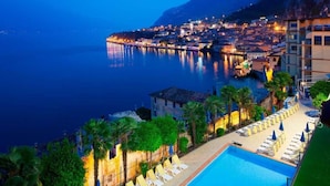 A coastal hotel with a swimming pool, palm trees, and a view of the city at night. - Hotel Splendid Palace (Limone sul Garda)