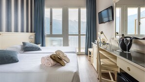 A hotel room with a bed, a desk, a TV, and a view of mountains through large windows. - Hotel Splendid Palace (Limone sul Garda)