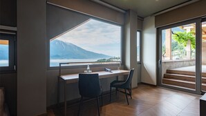 A modern room with a large window offering a view of a mountain and lake. There is a desk with two chairs and a phone. - Hotel Splendid Palace (Limone sul Garda)