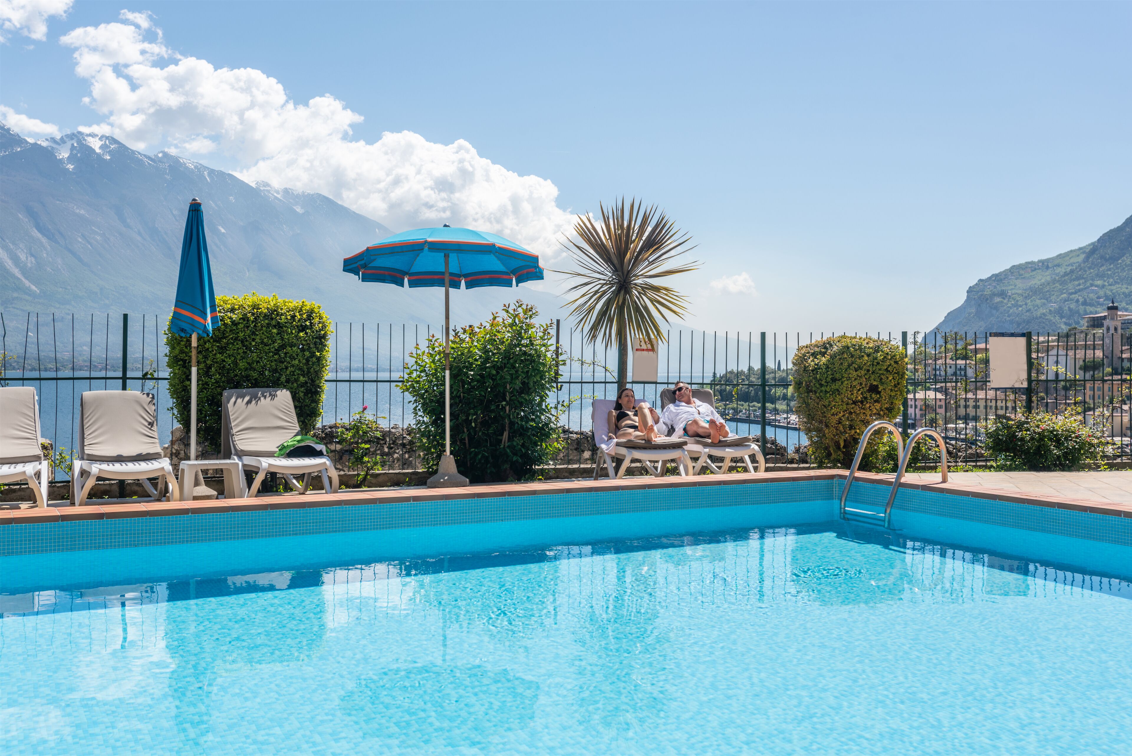 A swimming pool with lounge chairs and umbrellas, surrounded by a fence and greenery, with a mountain view in the background.