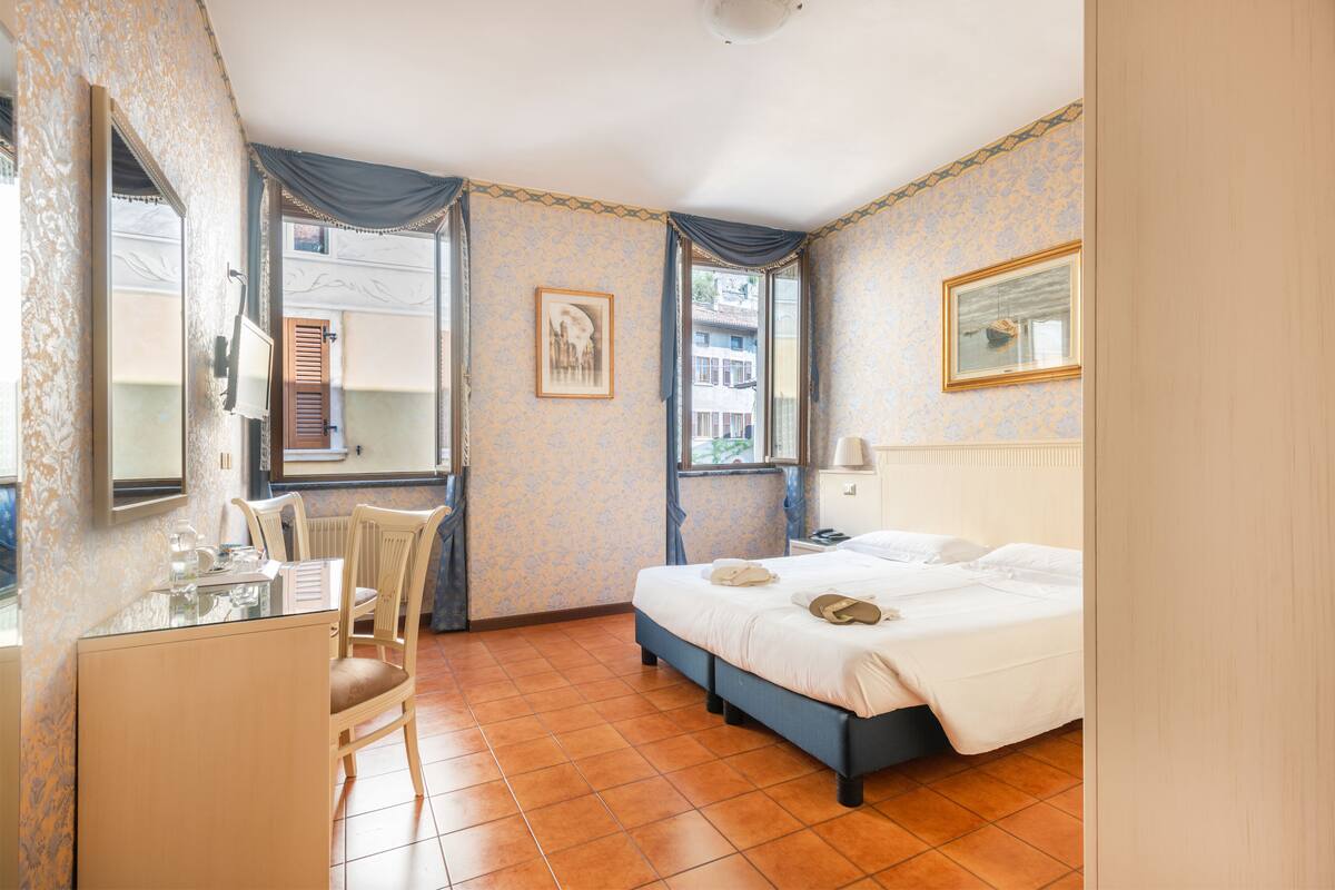 Ein Hotelzimmer mit Bett, Schreibtisch, Stuhl, einem Fenster mit Vorhängen und einem gerahmten Bild an der Wand.