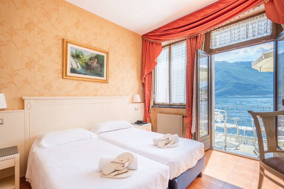 Ein Hotelzimmer mit zwei Betten, einem Bild an der Wand, Blick auf Meer und Berge durch ein großes Fenster und einem Sessel am Fenster.