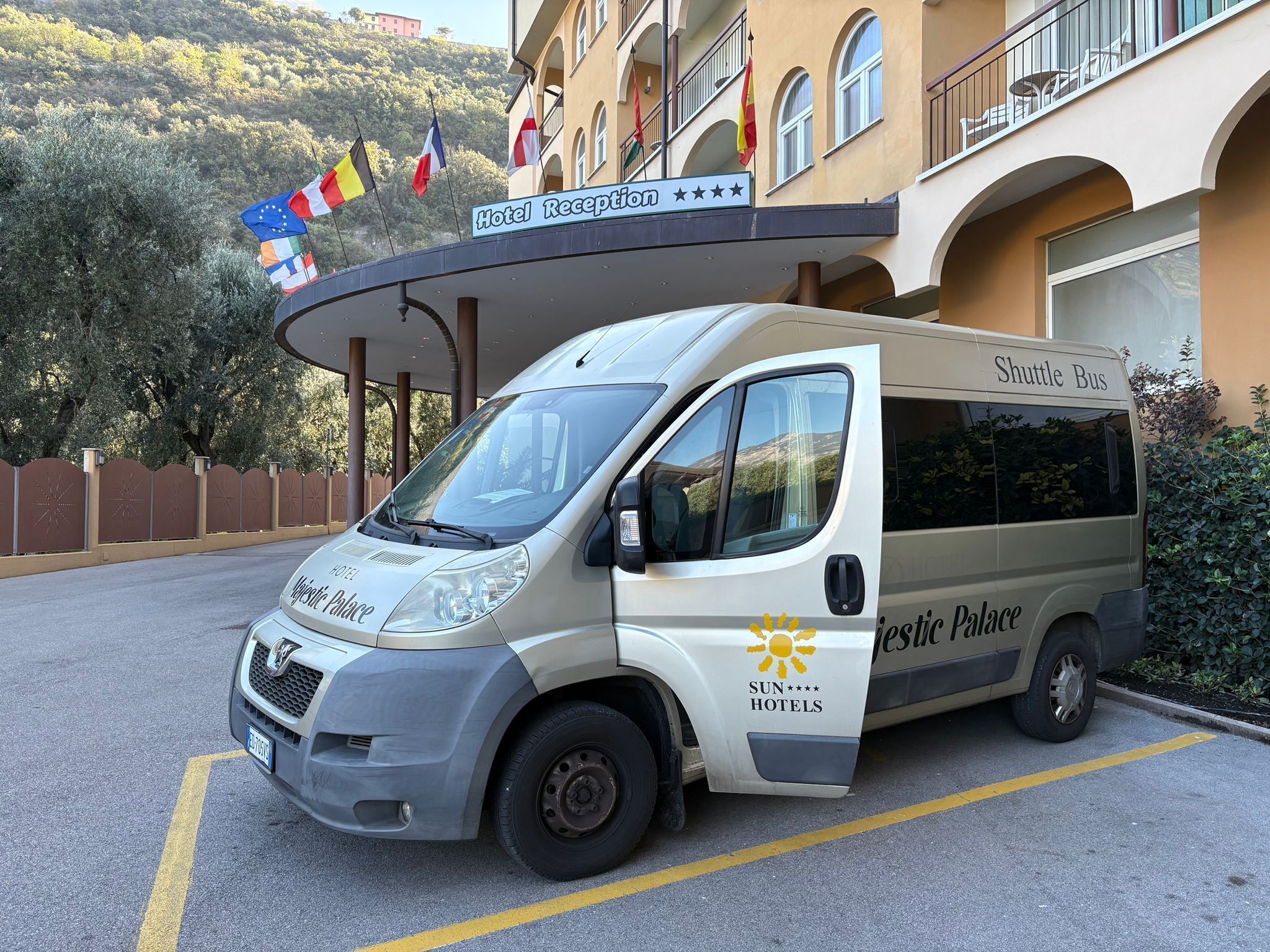 A shuttle bus parked in front of a hotel with flags, a reception area, and a building with balconies.