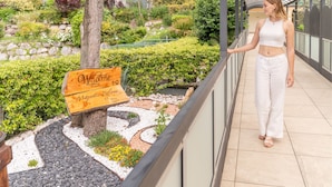 Une femme se promène sur un balcon d’où l’on peut admirer un jardin aménagé et une pancarte en bois sur laquelle on peut lire : « Bienvenue à Majestic Place ».