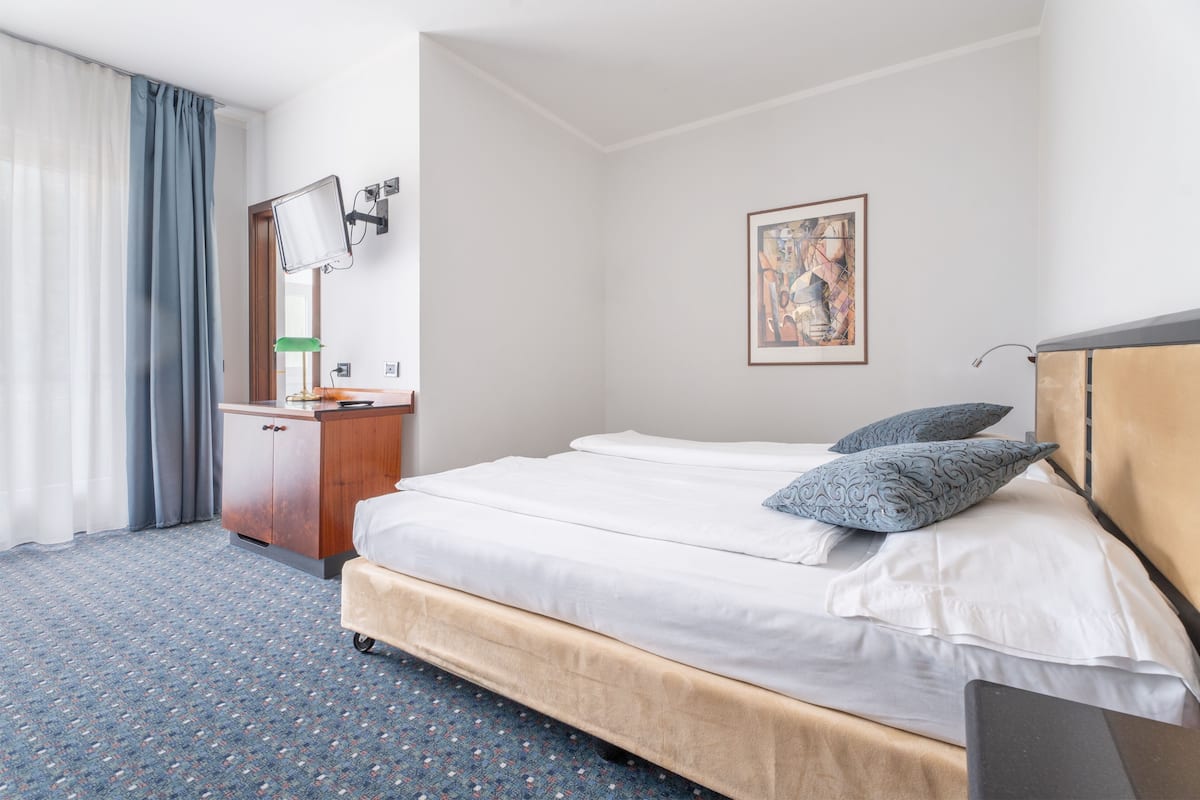 Une chambre d’hôtel avec un lit, un bureau, un miroir, un tableau au mur et une fenêtre avec des rideaux.