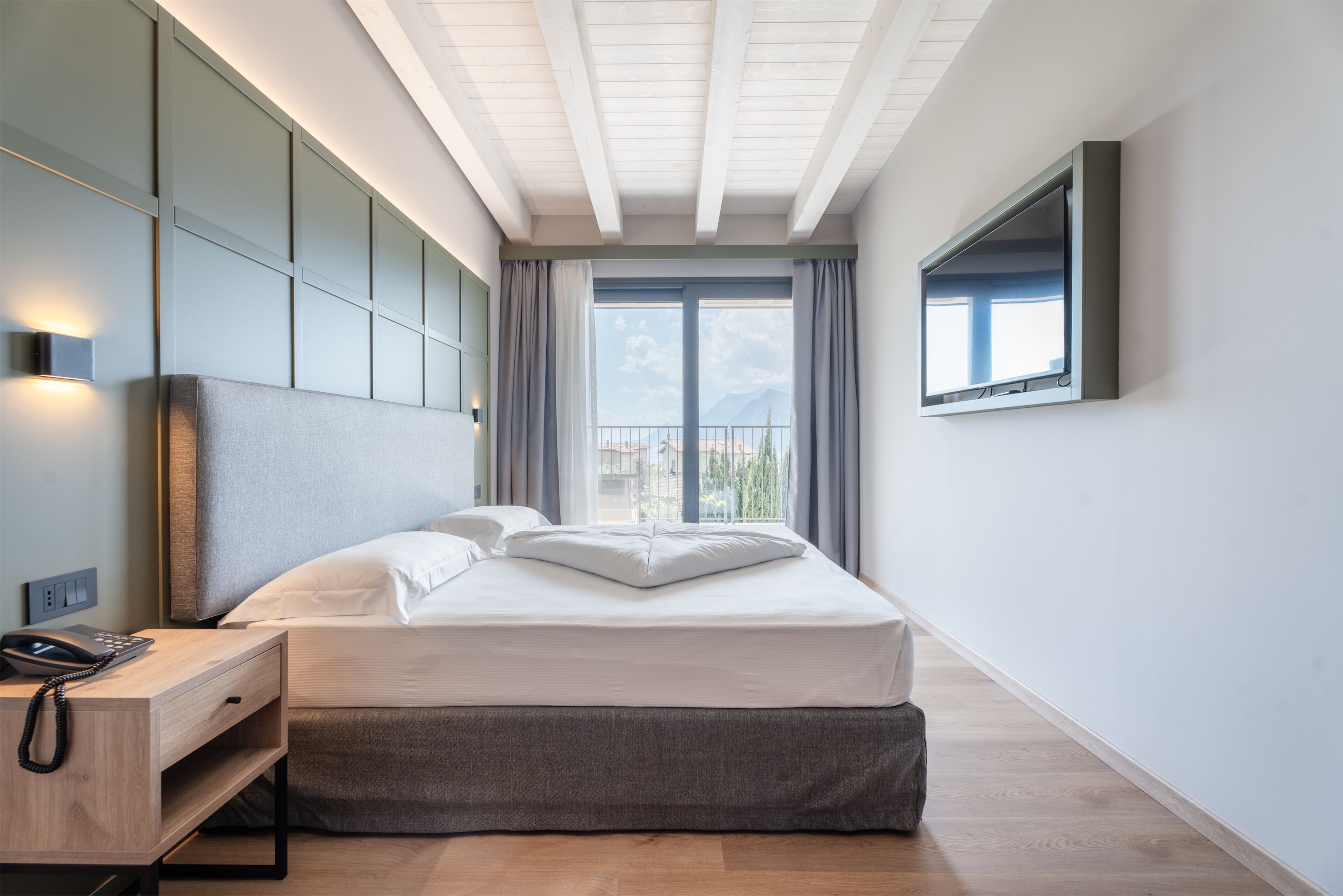 A modern hotel room with a large bed, a bedside table with a phone, a wall-mounted TV, and a view of the outdoors through a large window.