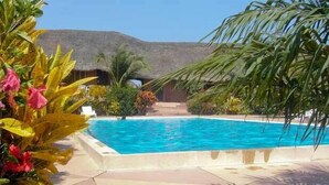 A swimming pool surrounded by tropical plants and a thatched-roof structure. - Hotel Maya (Cap Skirring)