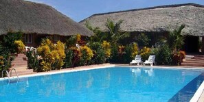 A swimming pool with lounge chairs, surrounded by tropical plants and thatched-roof buildings. - Hotel Maya (Cap Skirring)