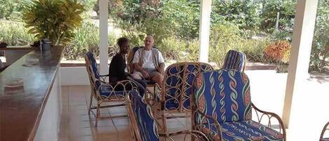 A covered outdoor seating area with wicker chairs and a long table, surrounded by lush greenery.