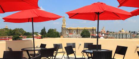 A terrace with outdoor seating and red umbrellas, overlooking a historic building with a clock tower.