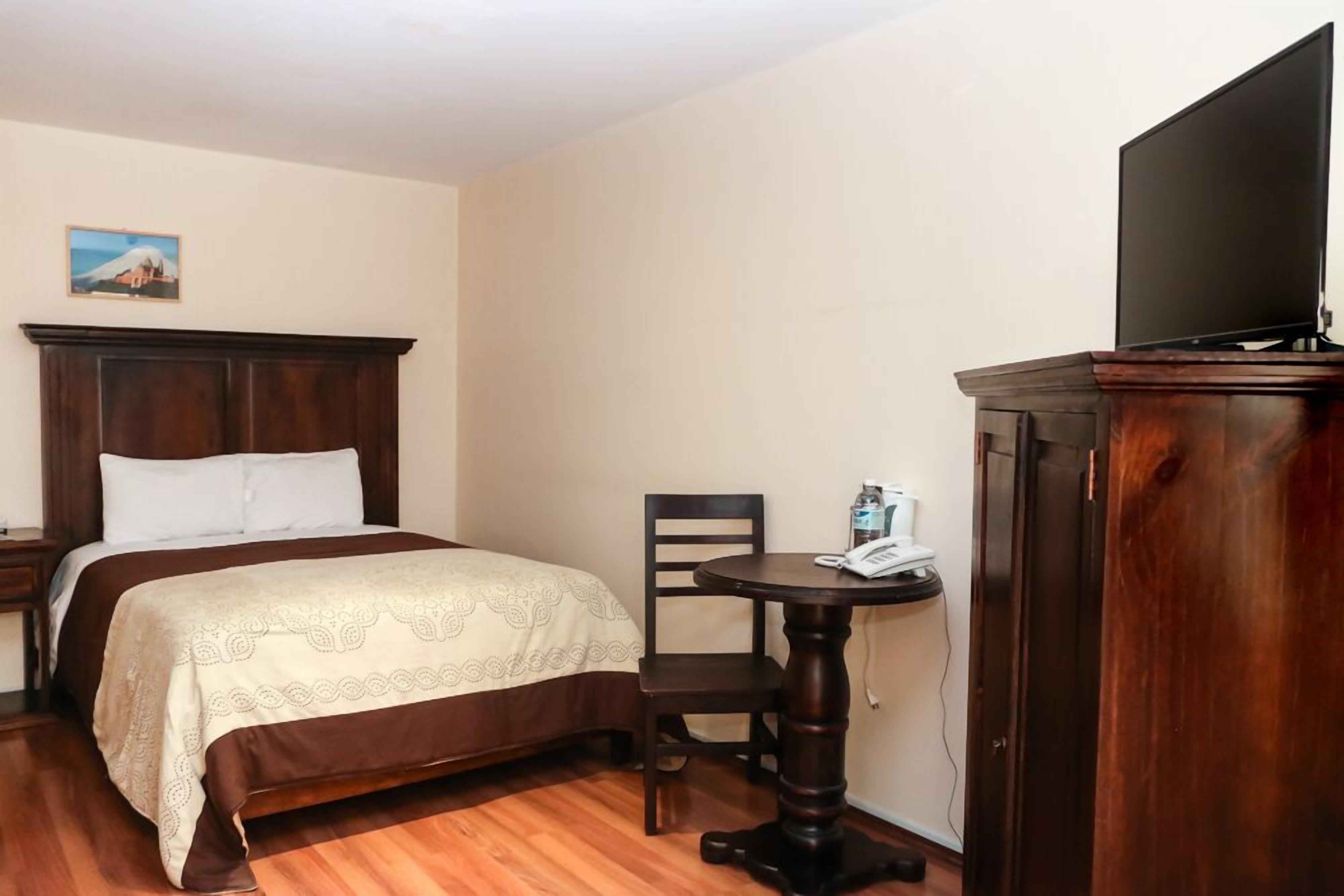 Una habitación de hotel con una cama, una mesita de noche de madera, una pequeña mesa redonda con una silla, un televisor sobre un mueble y un cuadro en la pared.
