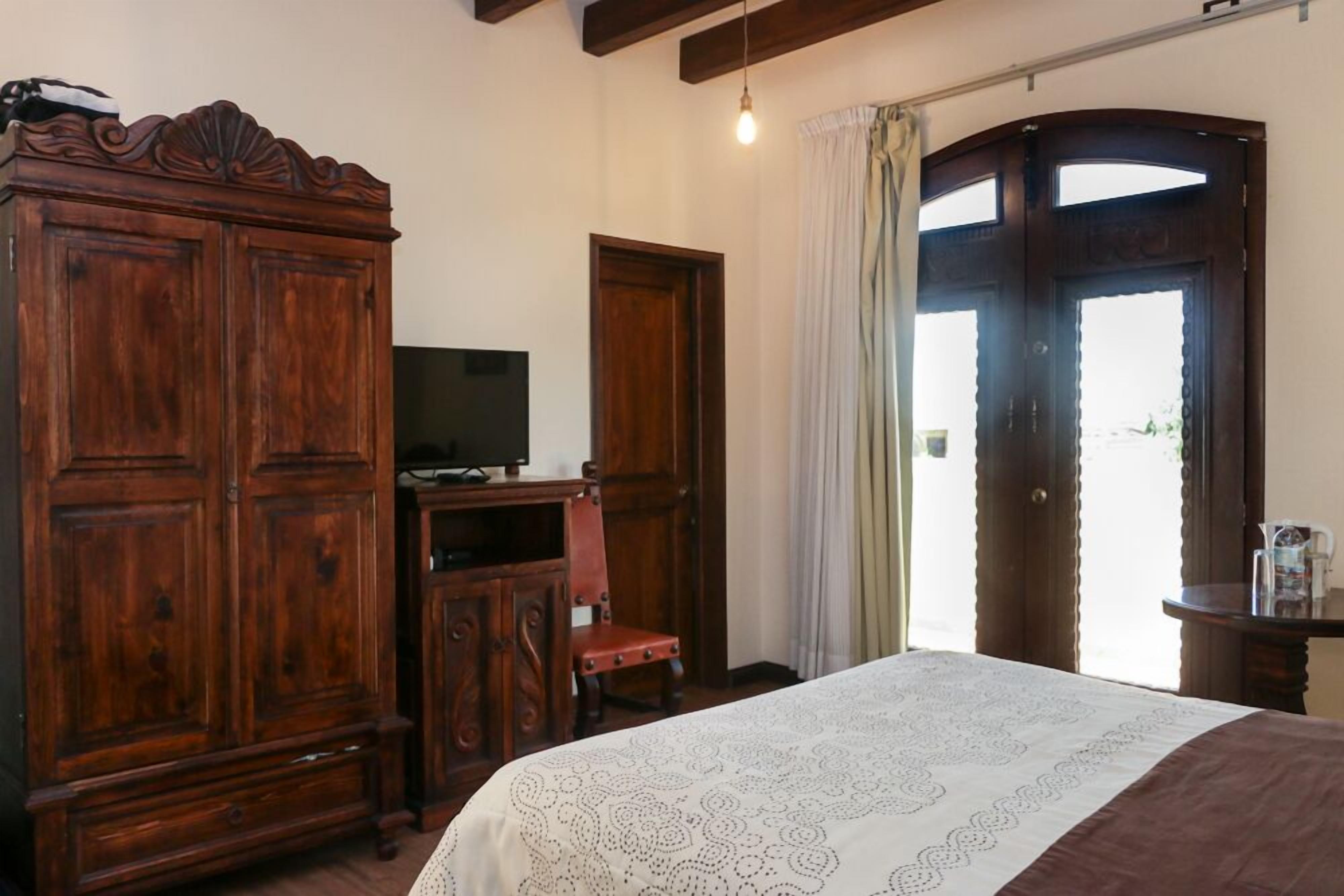 Une pièce comprenant une grande armoire en bois, un téléviseur posé sur un meuble en bois, un lit recouvert d’une couvre-lit à motifs et une porte avec des rideaux.