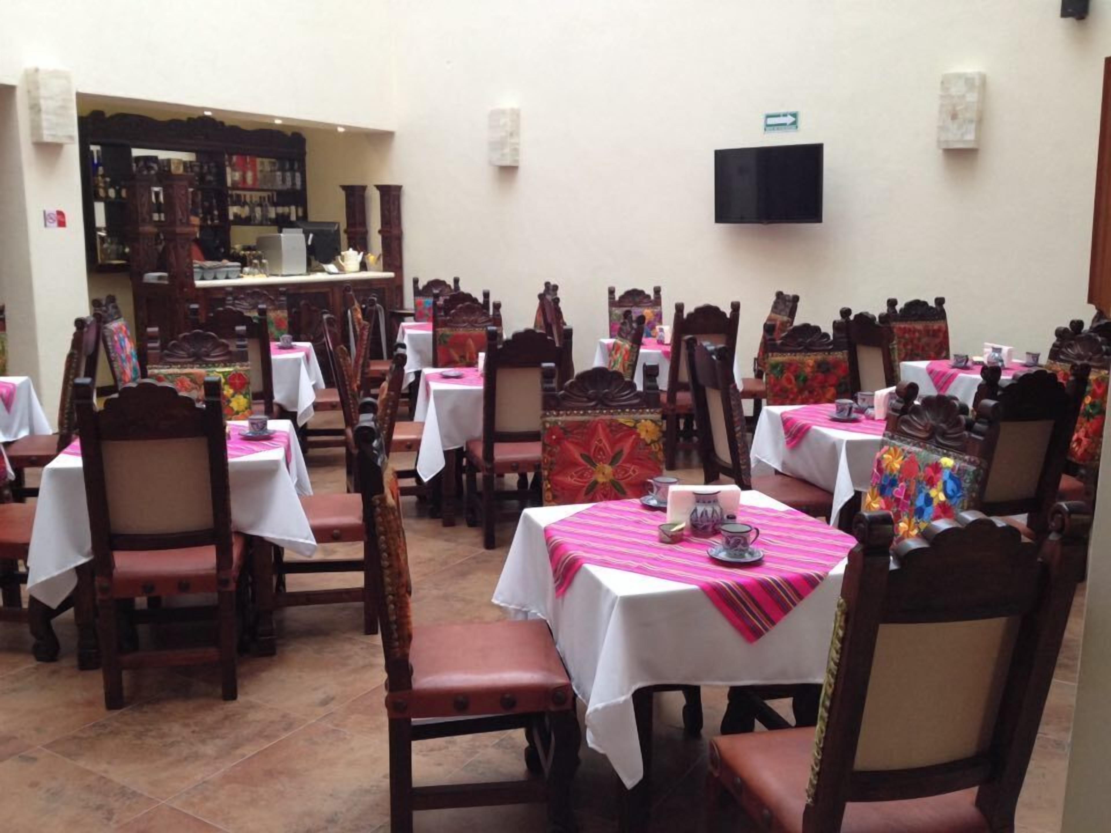 A restaurant with multiple tables and chairs, each with a white tablecloth and a pink table runner. There is a television mounted on the wall and a display case with various items in the background.