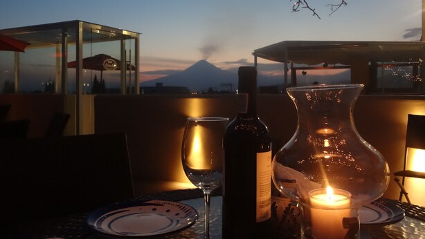 A table with a candle, a wine bottle, and a glass. In the background, there is a mountain and a clear sky. - Hotel Xelhua (San Pedro Cholula)