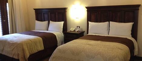 Two double beds with wooden headboards and white bedding, a bedside table with a lamp, and a window with curtains.