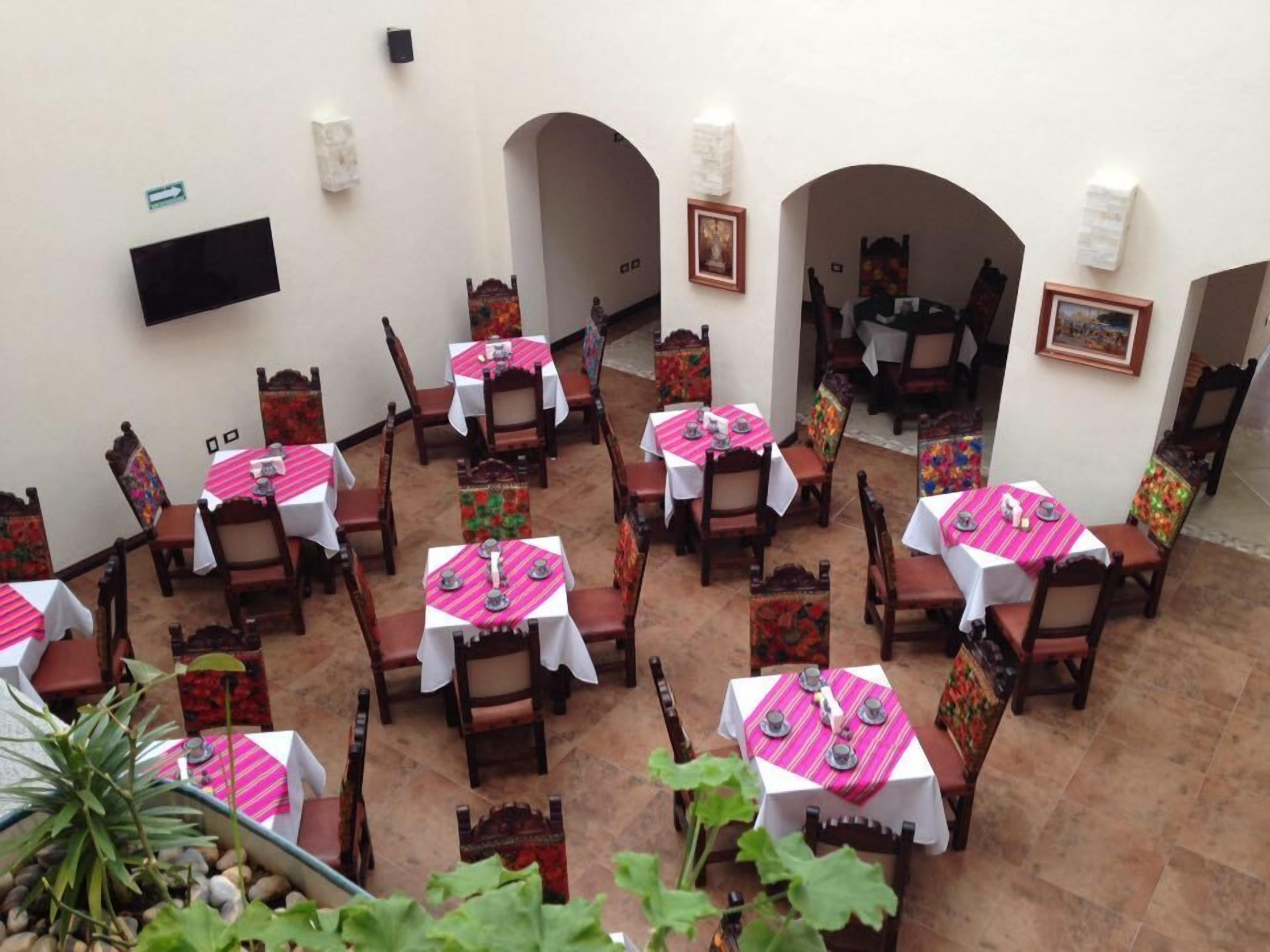Un restaurante con varias mesas, cada una cubierta con un mantel blanco y camino de mesa a rayas rosas. Las mesas están rodeadas de sillas de madera con asientos acolchados. El restaurante tiene arcos y un suelo de baldosas.