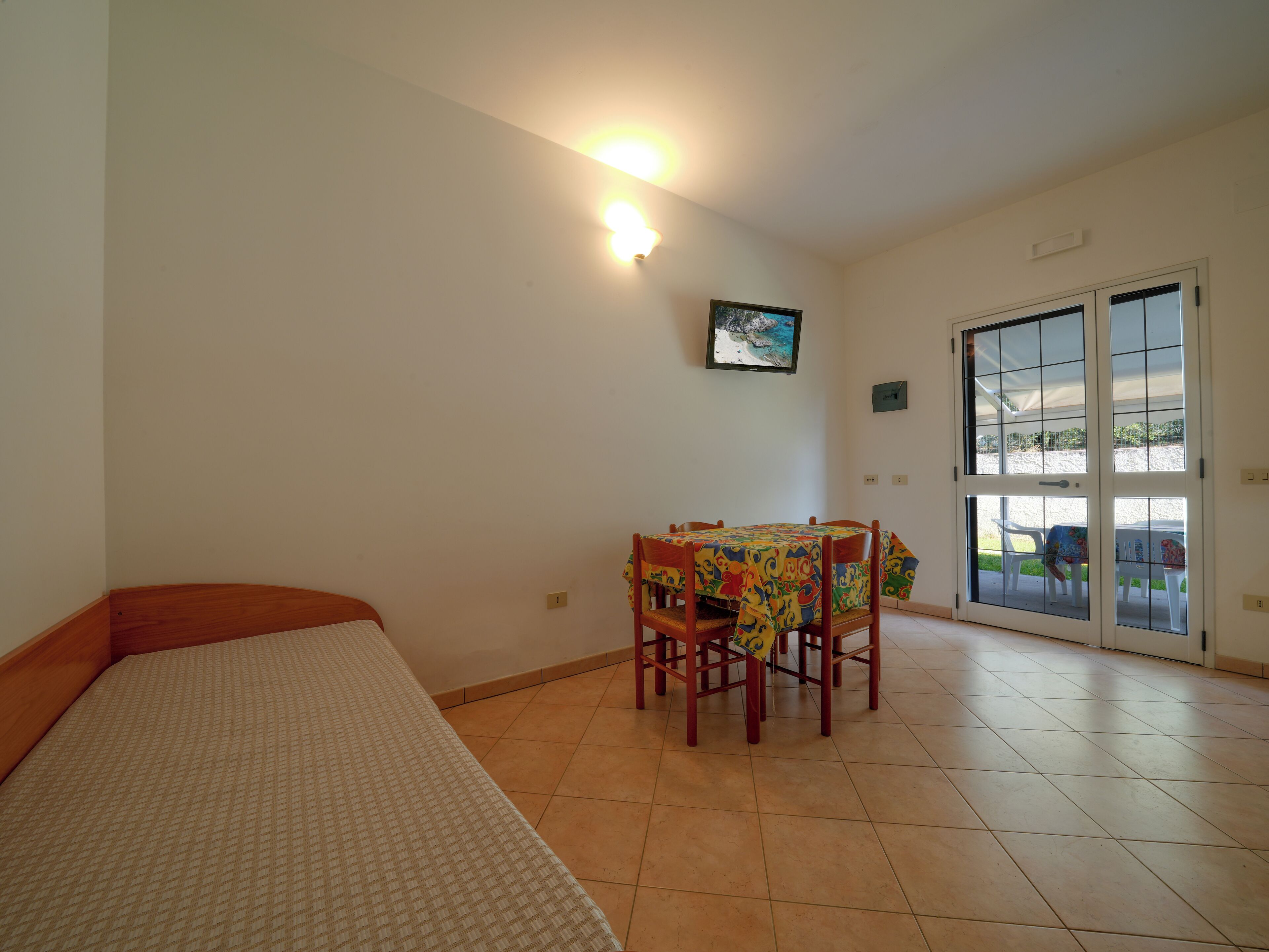 A room with a bed, a dining table with chairs, a television, and a sliding glass door leading to an outdoor area.
