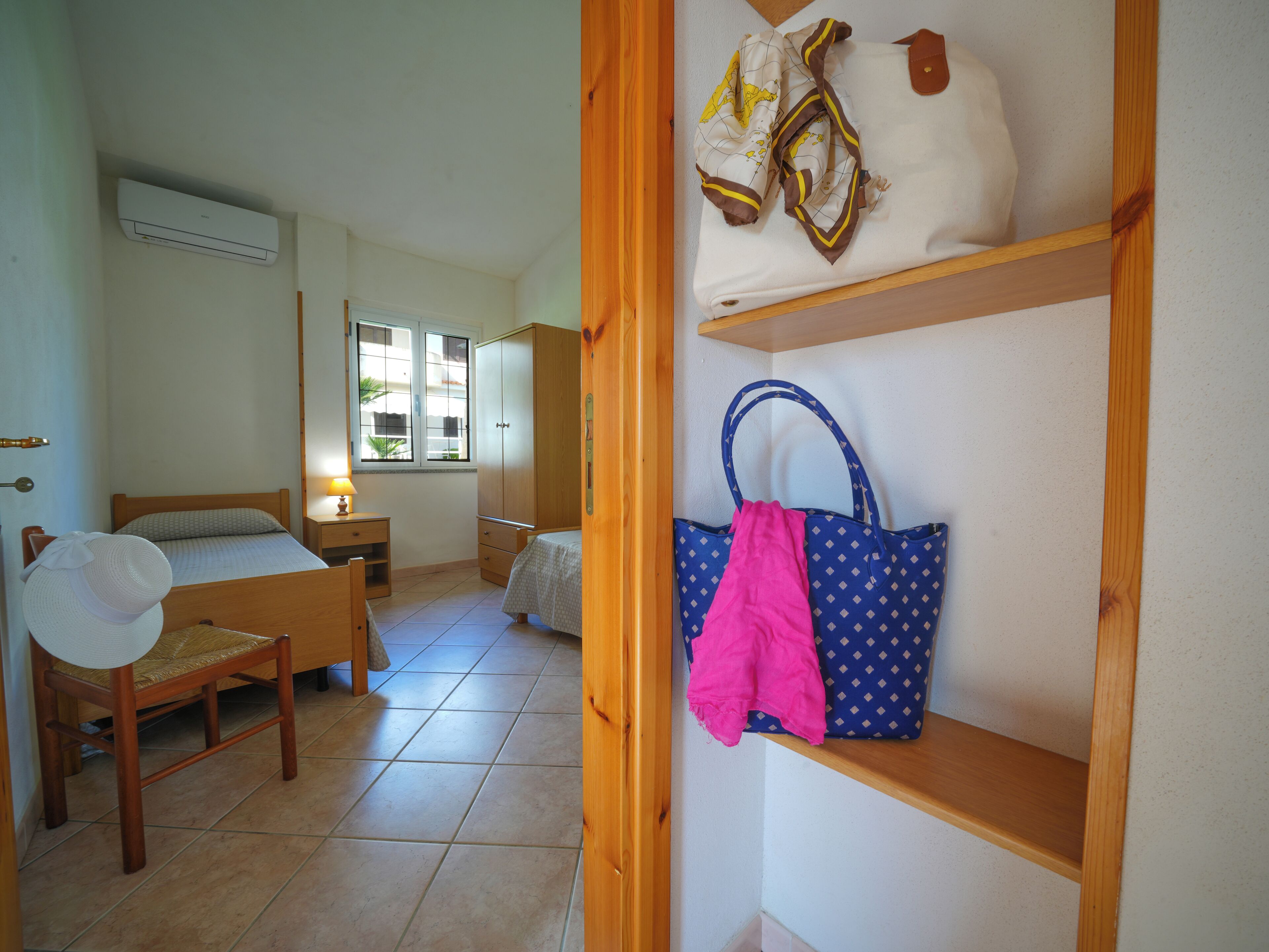 A bedroom with a bed, a chair, a nightstand, a wardrobe, and a shelf with a bag and a hat.